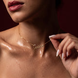 Woman wearing a gold-plated "Jessica" custom name necklace, holding the chain; soft light emphasizes her collarbone.