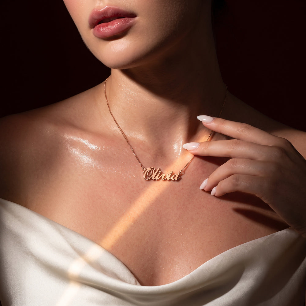 A close-up lifestyle shot of a woman wearing a delicate rose gold script necklace with the name 'Oliwia' on a dark background.
