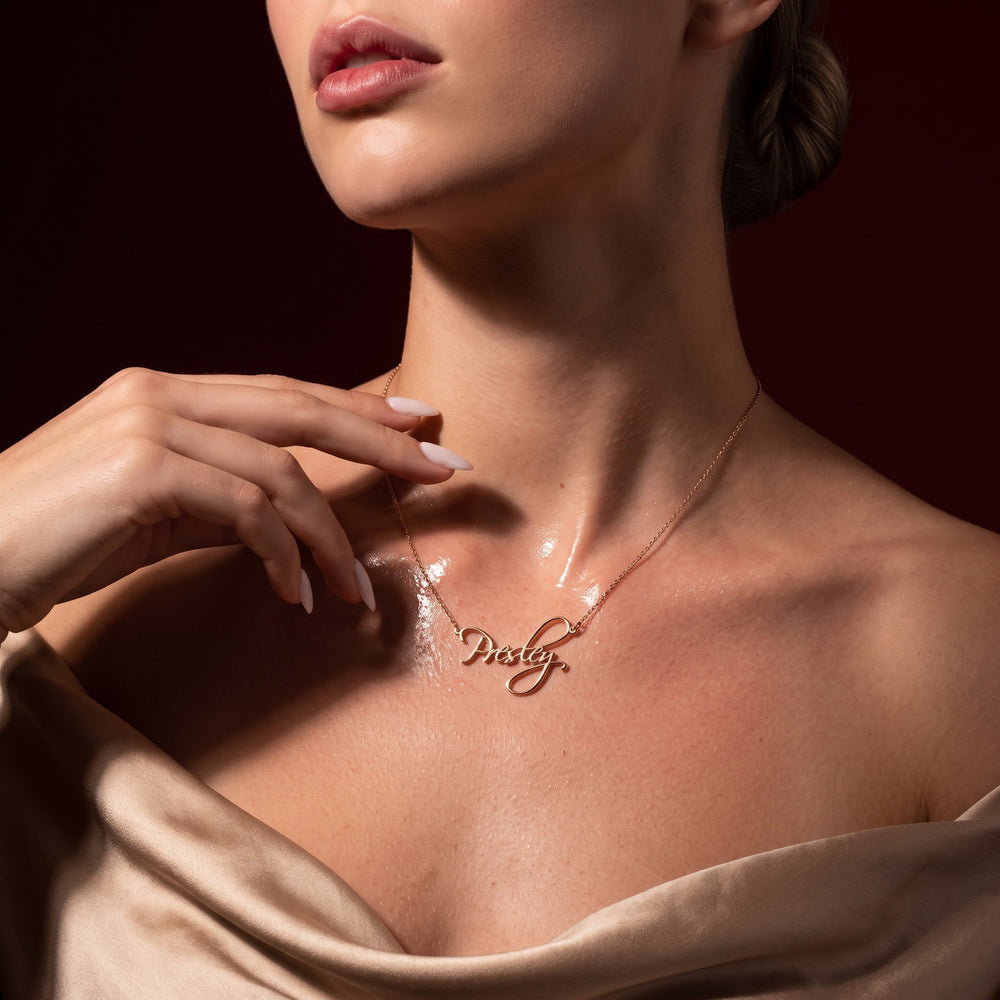 A close-up lifestyle shot of a model wearing a delicate rose gold script pendant necklace that spells 'Daisy'. The necklace is shown on the model's neck, with her hand resting near her shoulder, wearing a champagne satin top against a dark, warm brown background.